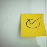Yellow sticky note with a check mark symbol on a white background, ideal for reminders.